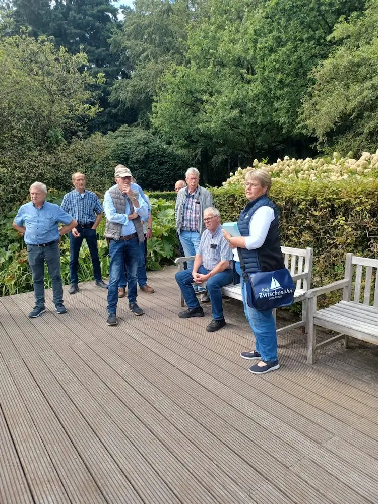 Eine Frau mit Infomaterial steht auf einer Terrasse in einem Garten und erklärt einer Gruppe Männer etwas