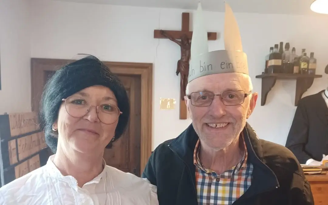 Eine Lehrerin vom "Damals" mit einem Herren, der eine Krone mit Ohren trägt, auf der "Ich bin ein Esel" steht.